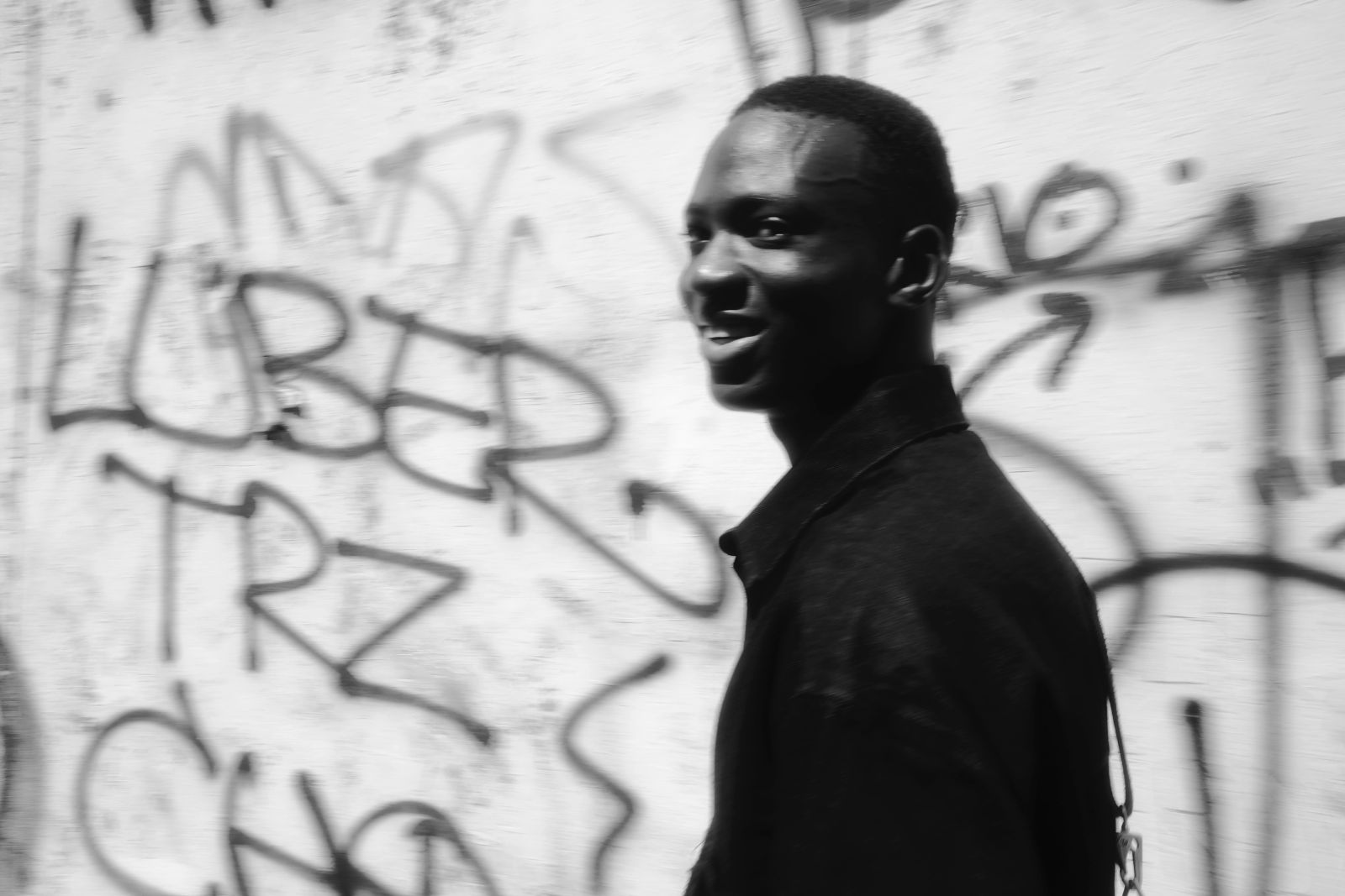 Schwarz-weiß Aufnahme mit künstlerischer Bewegungsunschärfe. Der junge Mann ist im Profil zu sehen, wie er lächelnd an einer Graffiti-Wand in Mailand vorbeigeht, was eine urbane Dynamik erzeugt, fotografiert von Karl Scholten