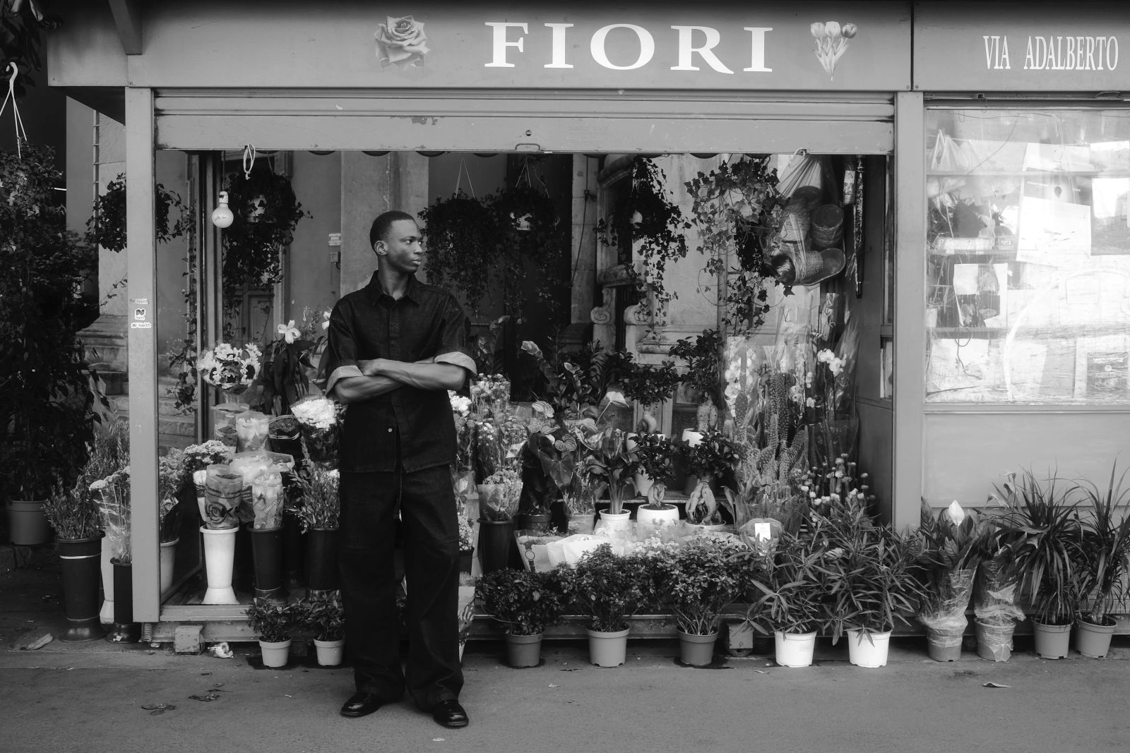 Weitwinklige schwarz-weiß Aufnahme. Der junge Mann steht mit verschränkten Armen vor einem traditionellen Mailänder Blumenladen mit dem Schild "FIORI" und "VIA ADALBERTO", umgeben von zahlreichen Topfpflanzen, fotografiert von Karl Scholten.