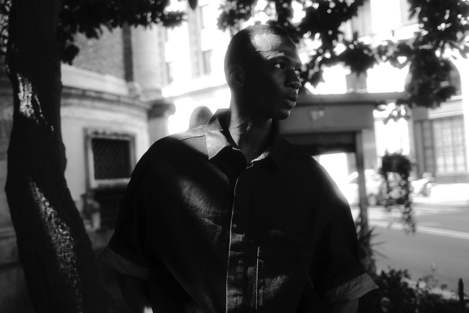Schwarz-weißes Außenporträt in Mailand. Der junge Mann steht unter Bäumen, wobei starkes, geflecktes Sonnenlicht (Chiaroscuro-Effekt) auf sein Gesicht und sein dunkles Hemd fällt. Im Hintergrund ist klassische Architektur zu erahnen fotografiert von Karl Scholten.