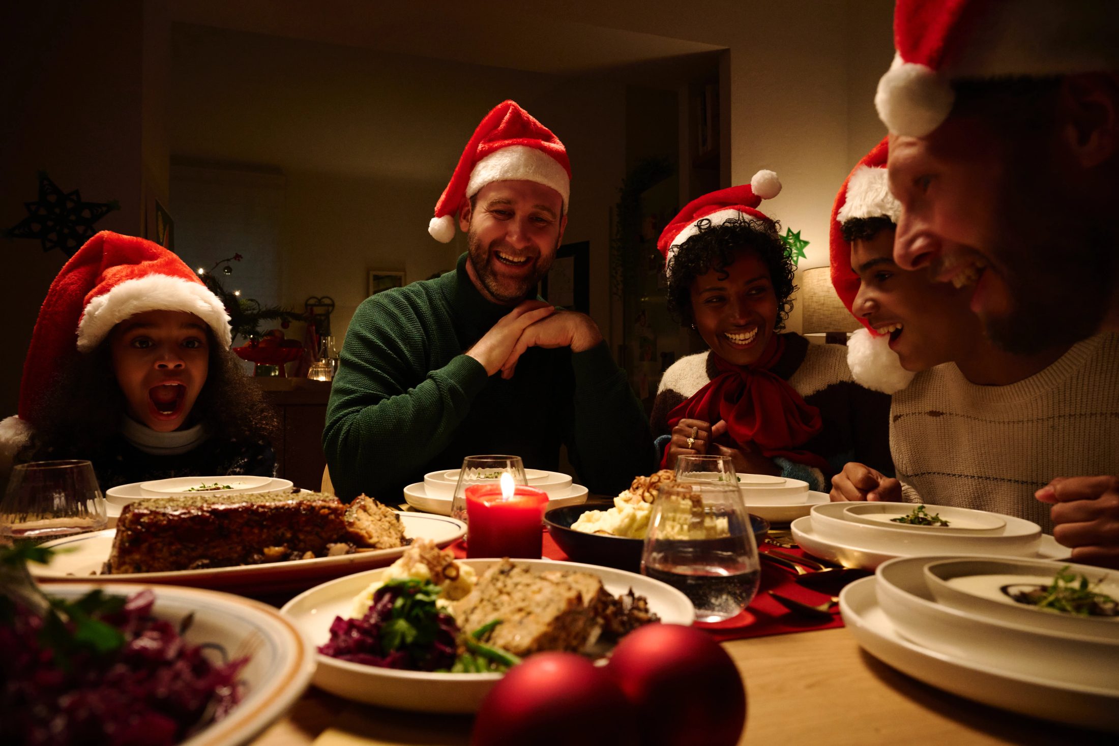 Mehrere Personen mit Weihnachtsmützen sitzen lachend an einer festlichen Tafel und genießen ihr Essen