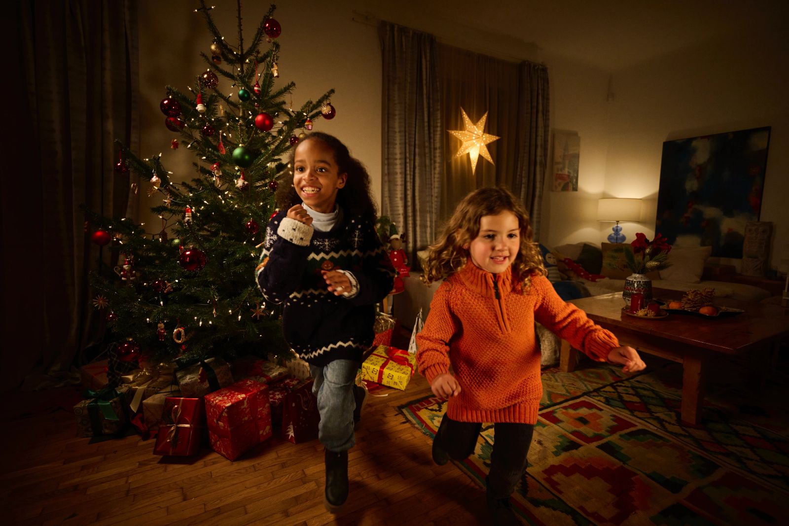 Zwei Personen laufen freudig durch ein weihnachtlich dekoriertes Wohnzimmer neben einem leuchtenden Tannenbaum