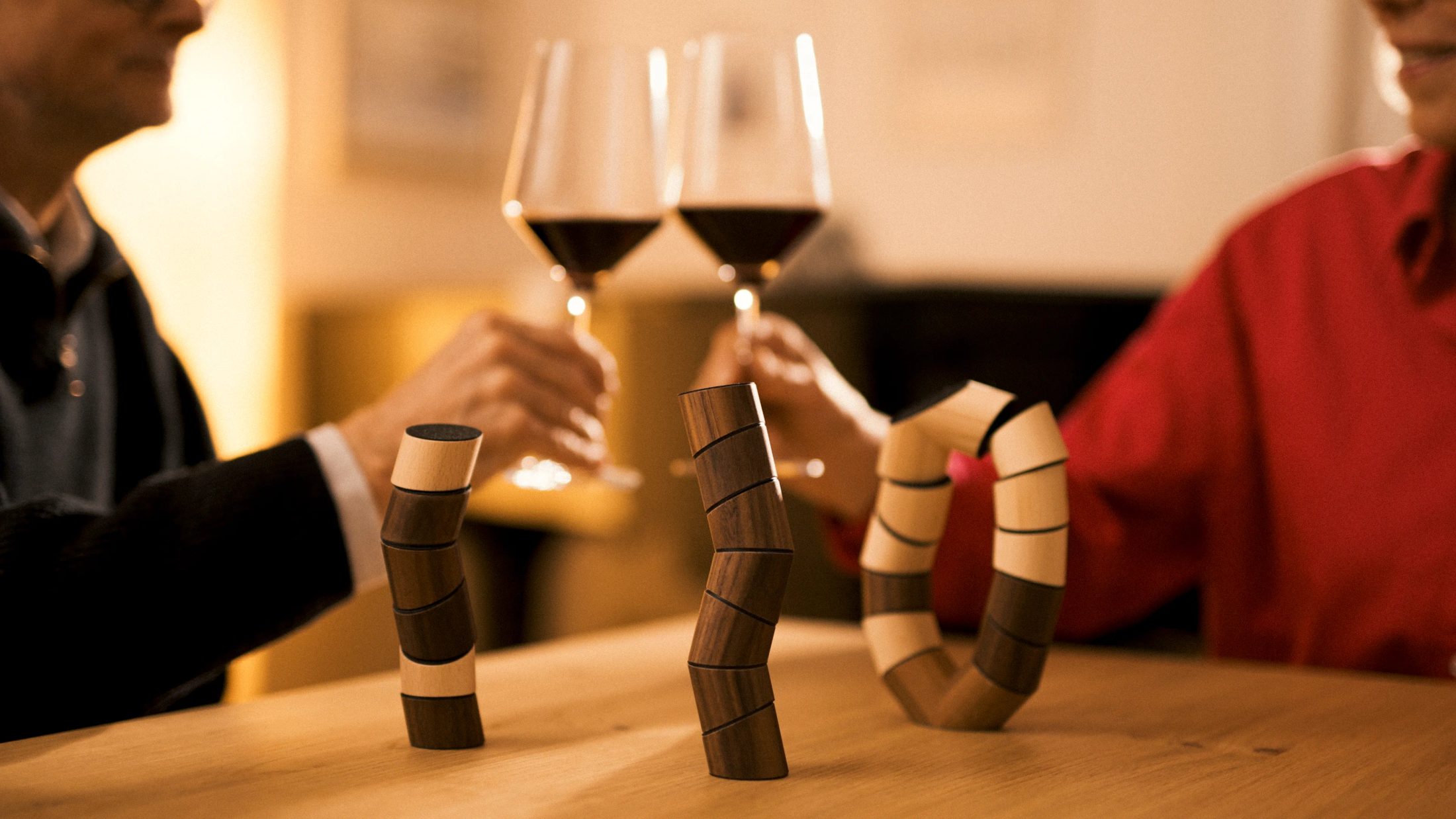 Lifestyle-Moment zu Hause: Drei fertige Holzspielzeug-Figuren stehen im Fokus auf einem Holztisch, während im Hintergrund zwei Personen anstoßen, fotografiert von Karl Scholten.