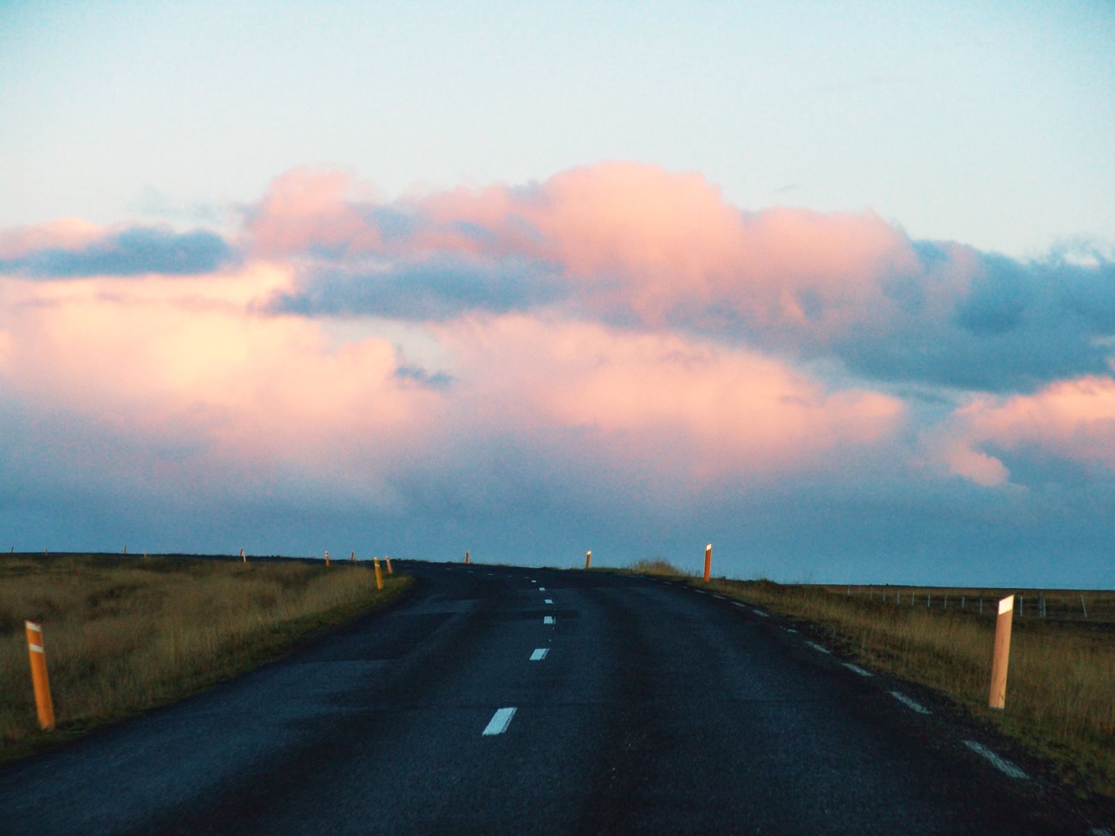 Eine leere, asphaltierte Straße mit gelben Begrenzungspfählen führt in Island unter rosa leuchtenden Abendwolken über eine Kuppe, fotografiert von Karl Scholten.