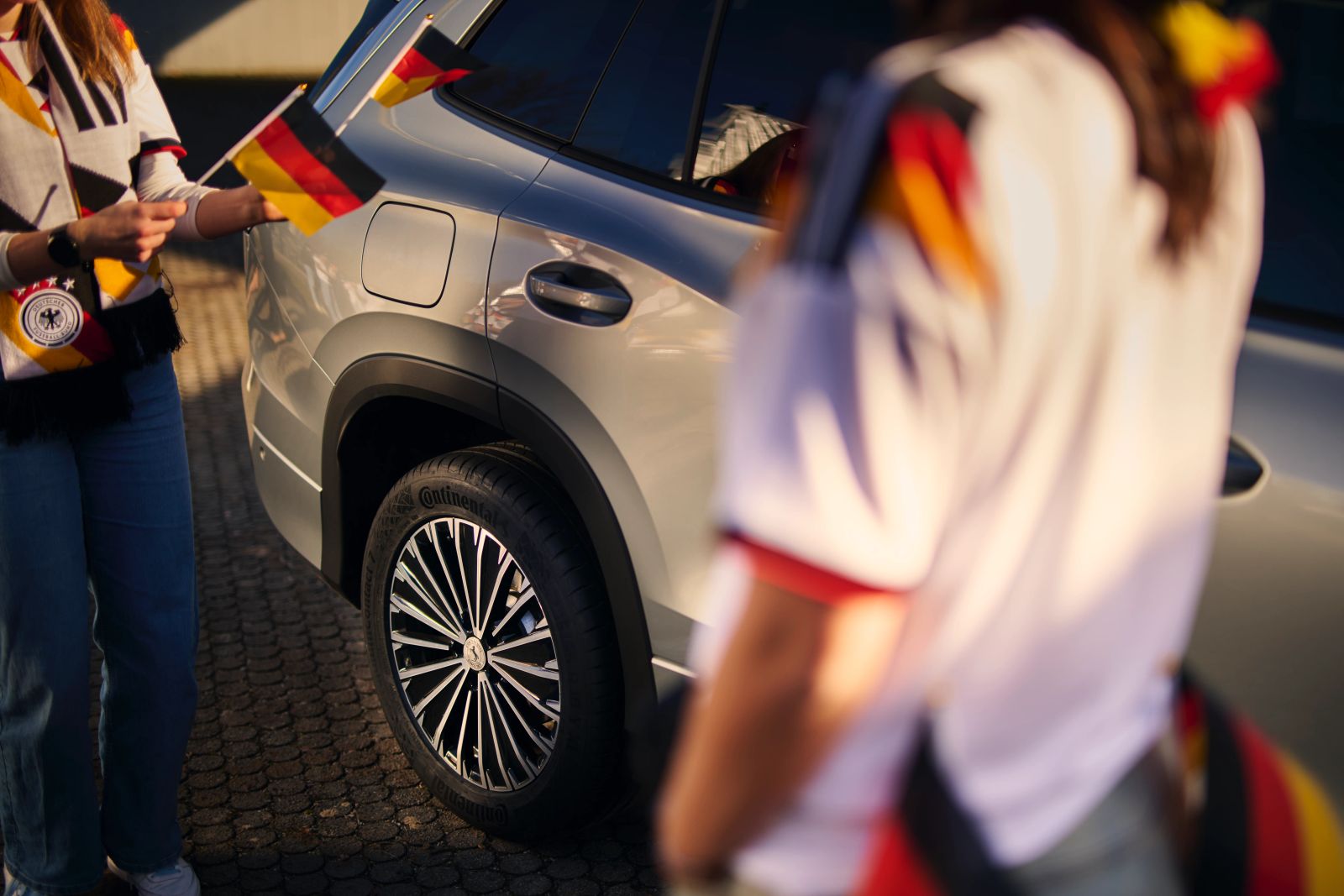 Zwei ausgelassene Fans in Deutschland-Trikots und mit Fanutensilien jubeln vor einem silbernen Continental-Fahrzeug; Werbemotiv für die Continental WM 2026 Kampagne, fotografiert von Karl Scholten