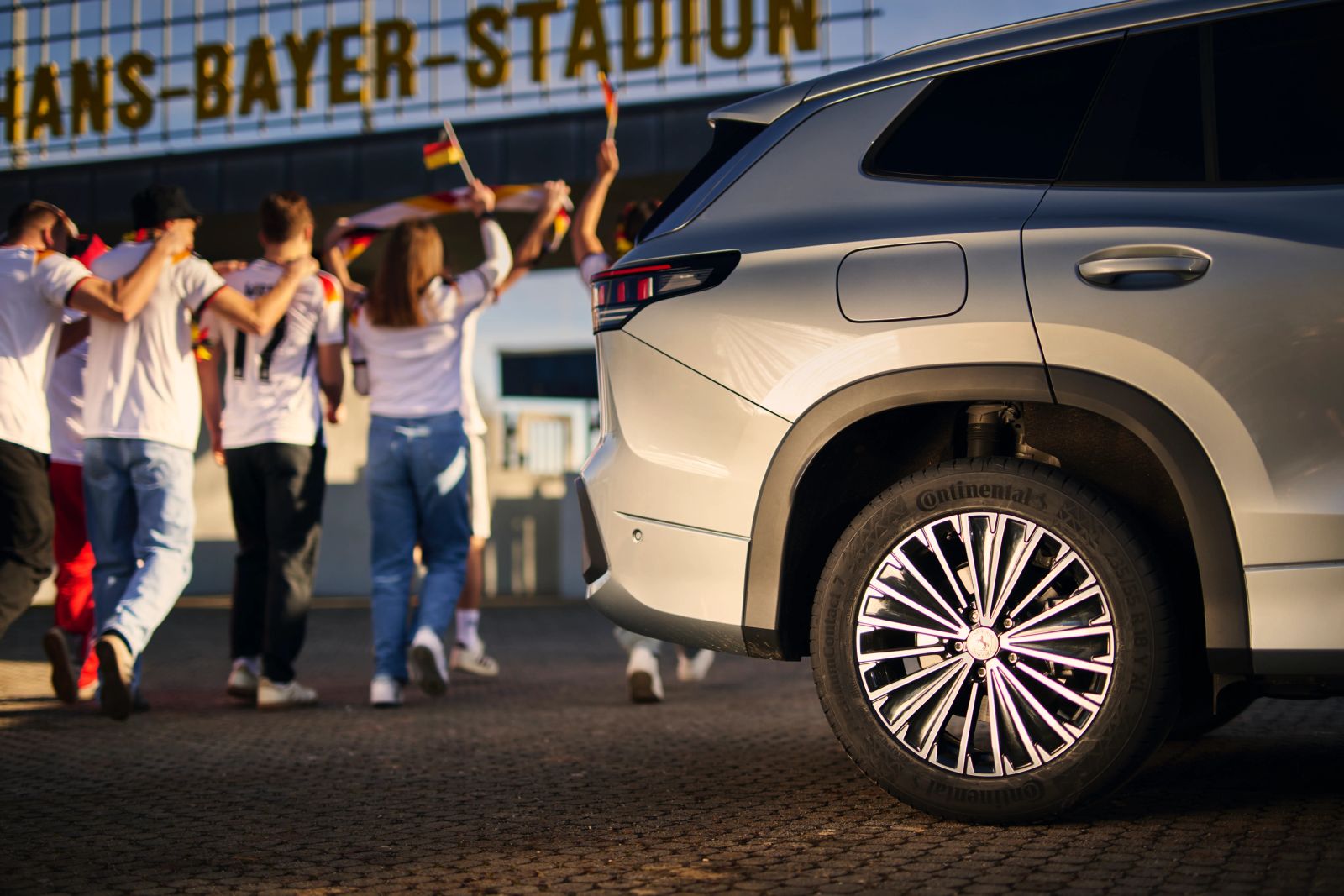 Seitenansicht eines silbernen VW-SUVs mit Continental-Reifen vor dem Hans-Bayer-Stadion, im Hintergrund laufen jubelnde Fans