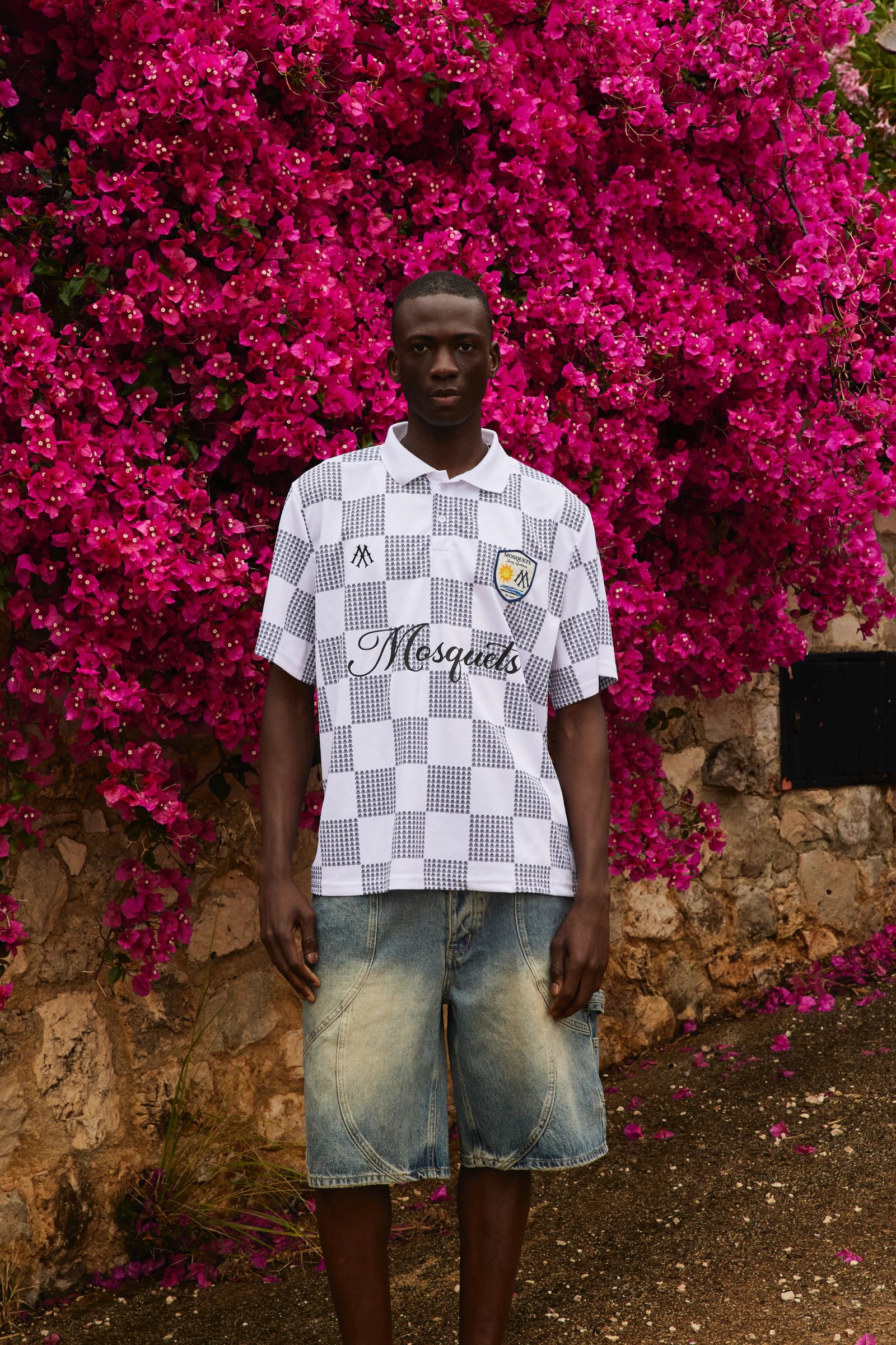 Mann in Mosquets-Poloshirt und Jeansshorts vor pinker Bougainvillea-Wand in Sizilien