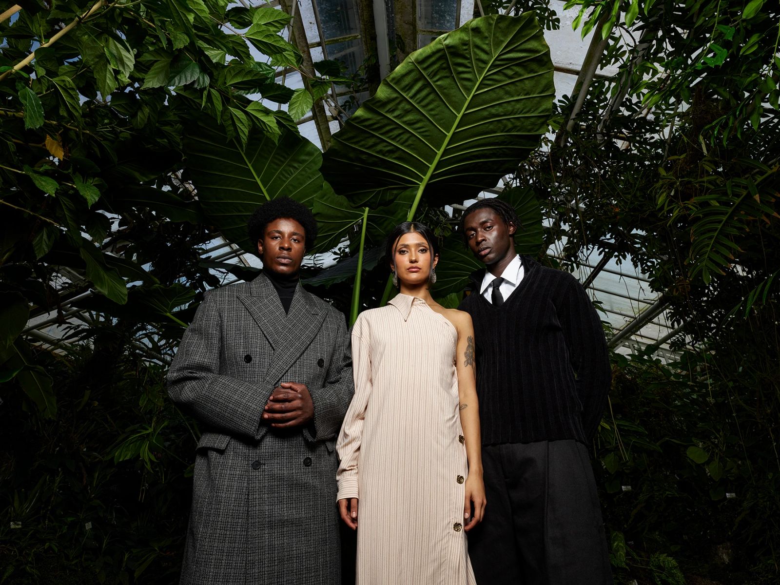 Drei Models in High-Fashion-Outfits, unter anderem ein cremefarbenes One-Shoulder-Kleid, stehen unter großen Tropenblättern in einem Glashaus