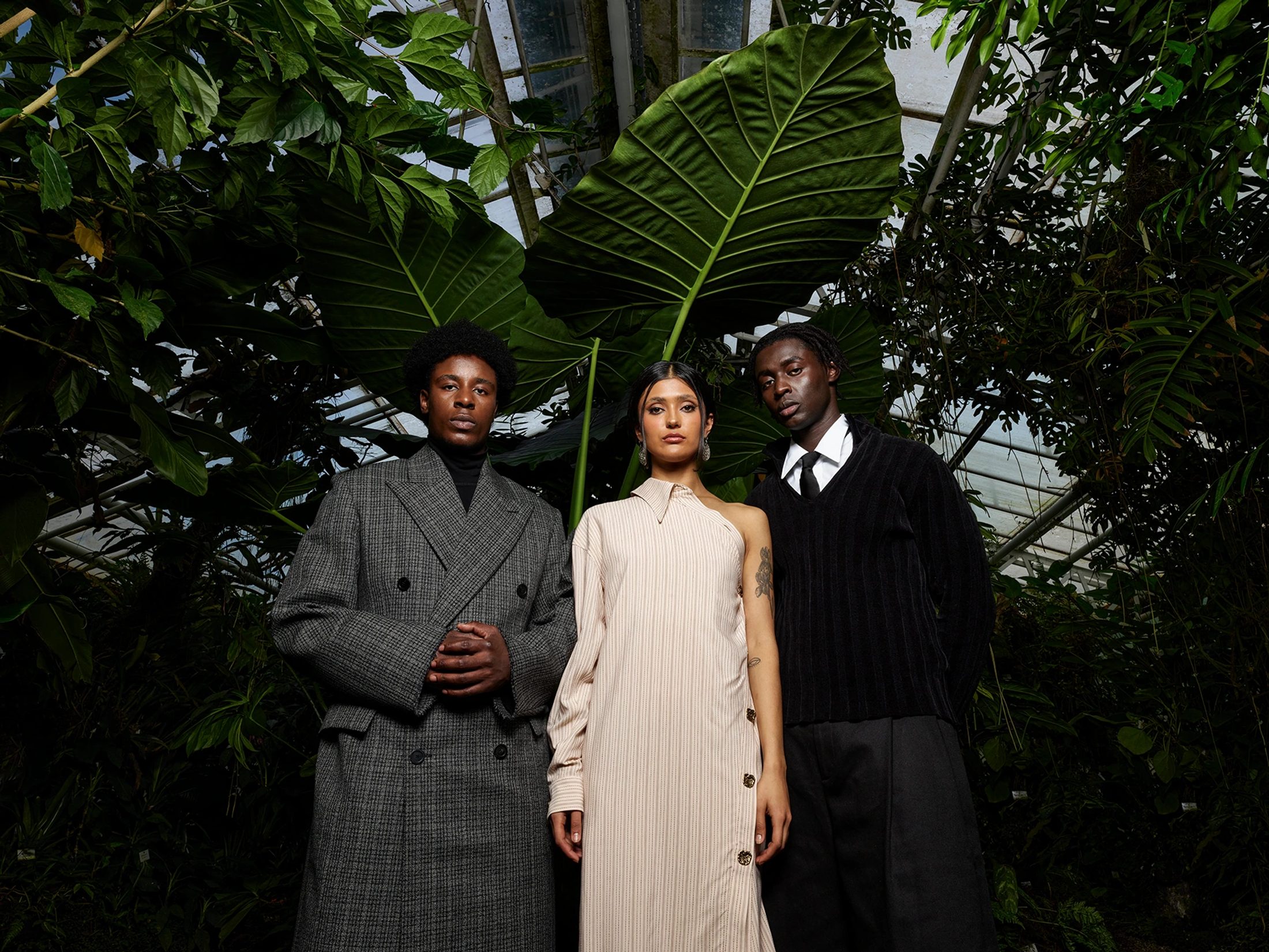 Drei Models in High-Fashion-Outfits, unter anderem ein cremefarbenes One-Shoulder-Kleid, stehen unter großen Tropenblättern in einem Glashaus
