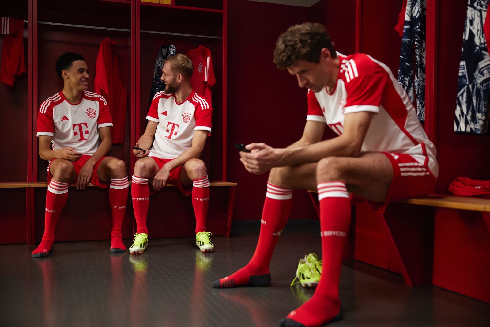 Thomas Müller am Handy, während Jamal Musiala und Konrad Laimer lachend in den Kabinen der Allianz Arena im Hintergrund sitzen, fotografiert von Karl Scholten.