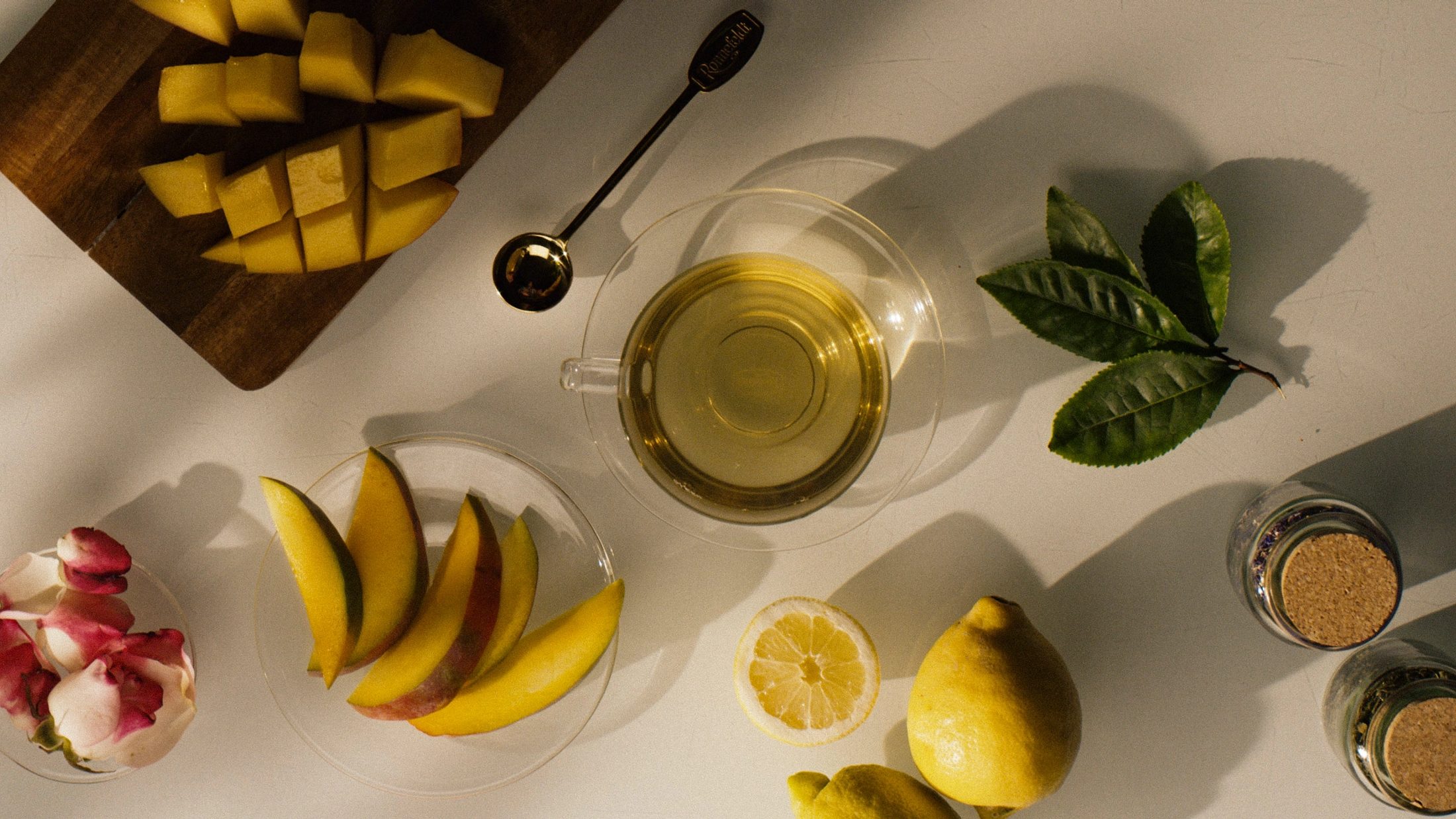 Vogelperspektive auf eine Tasse Tee, umgeben von frischen Mango-Stücken, Zitronen, Rosenblättern und Teeblättern auf einem hellen Tisch mit markantem Schattenwurf