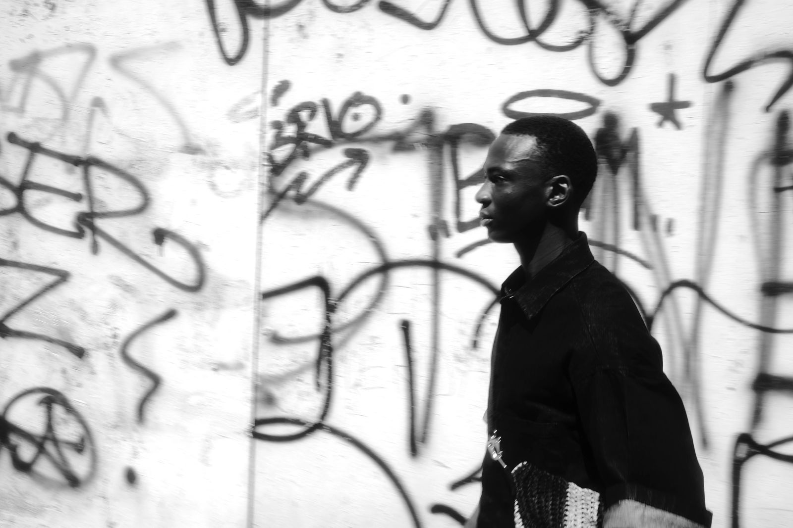 Schwarz-weißes Fashion-Foto. Eine klare Profilaufnahme des jungen Mannes mit Heiligenschein, der entschlossen an einer hell erleuchteten, weißen Wand vorbeigeht, die mit deutlichen schwarzen Graffiti-Tags versehen ist.