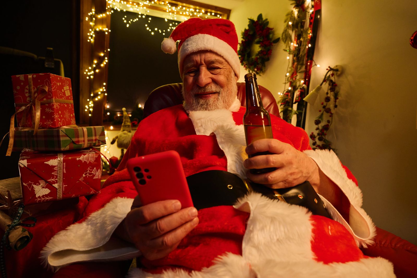 ine Person im Weihnachtsmann-Kostüm sitzt in einem Sessel, blickt auf ein Smartphone und hält eine Glasflasche in der Hand