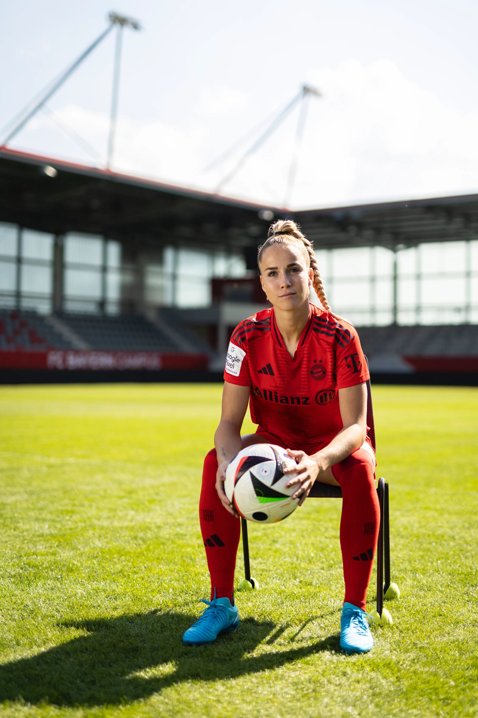 Frontales Porträt von FC Bayern Spielerin Giulia Gwinn, die mit einem Fußball auf einem Stuhl mitten auf dem Rasen sitzt, fotografiert von Karl Scholten.