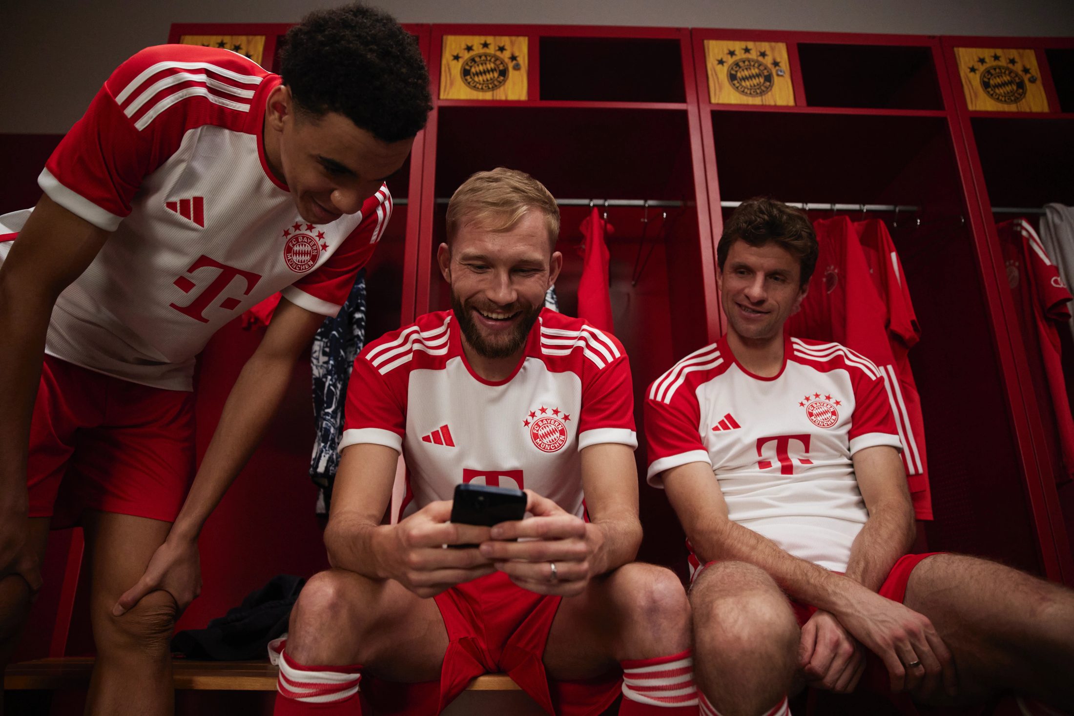 Jamal Musiala, Konrad Laimer und Thomas Müller blicken in den Kabinen der Allianz Arena gemeinsam auf ein Smartphone für Bitpanda, fotografiert von Karl Scholten.