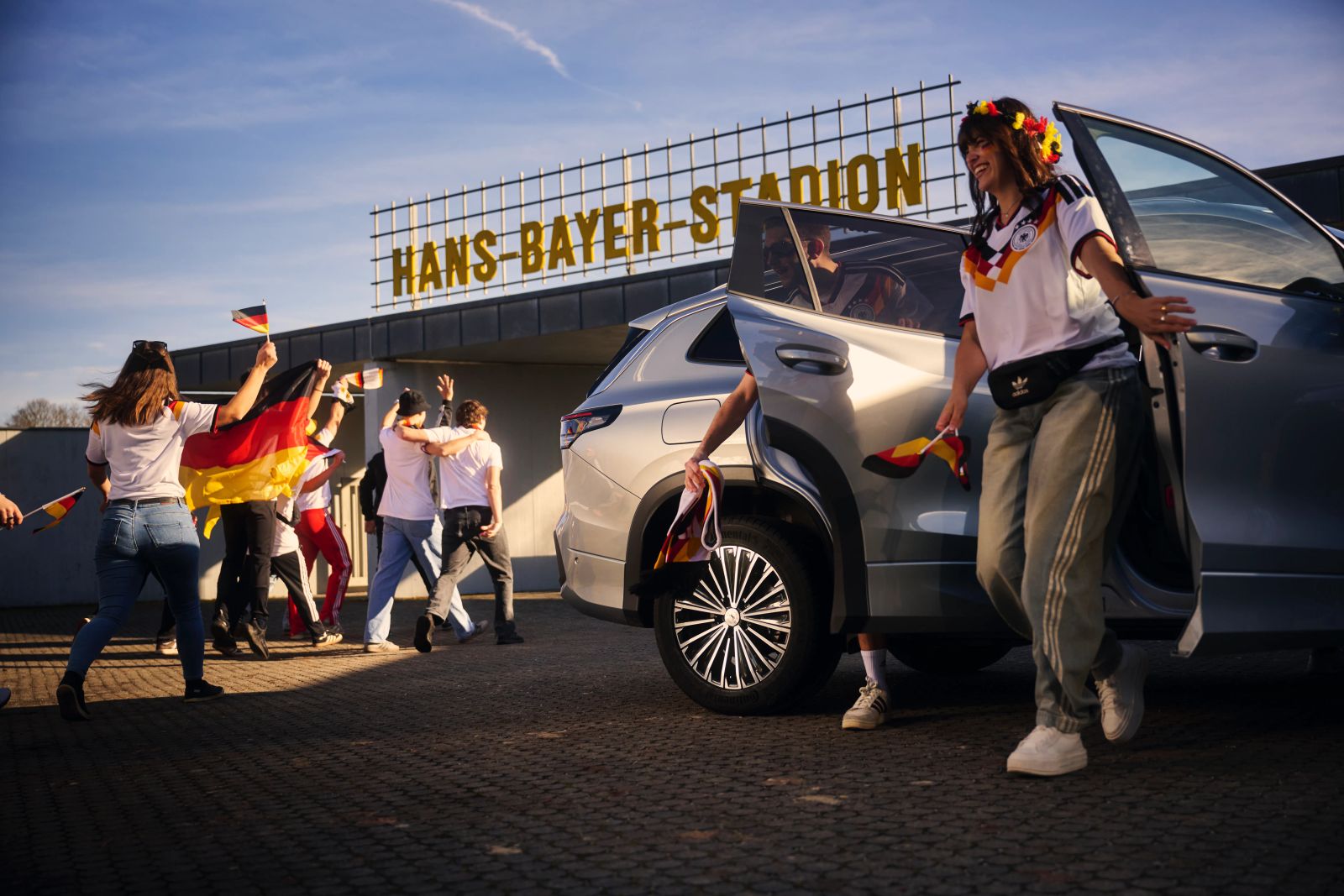Deutsche Fußballfans steigen gut gelaunt aus einem silbernen VW-SUV vor dem Hans-Bayer-Stadion aus.