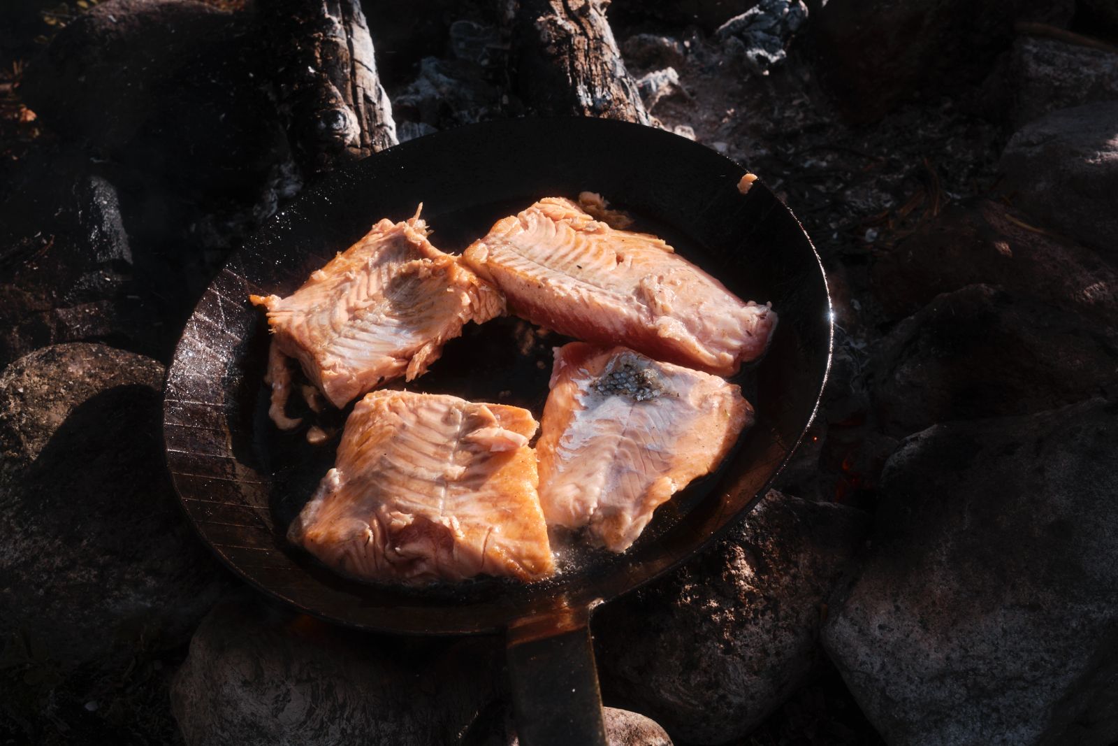 Fischfilets garen in einer Eisenpfanne über der Glut eines Campfeuers.