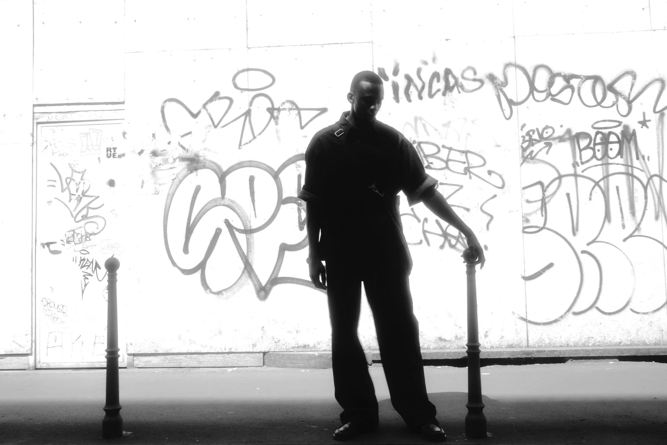 Schwarz-weißes Fashion-Editorial-Foto in Mailand. Ein junger Mann in dunkler, weiter Kleidung steht in einer Ganzkörperaufnahme vor einer stark mit Graffiti besprühten Wand und lehnt seine Hand lässig auf einen kleinen Metallpoller.