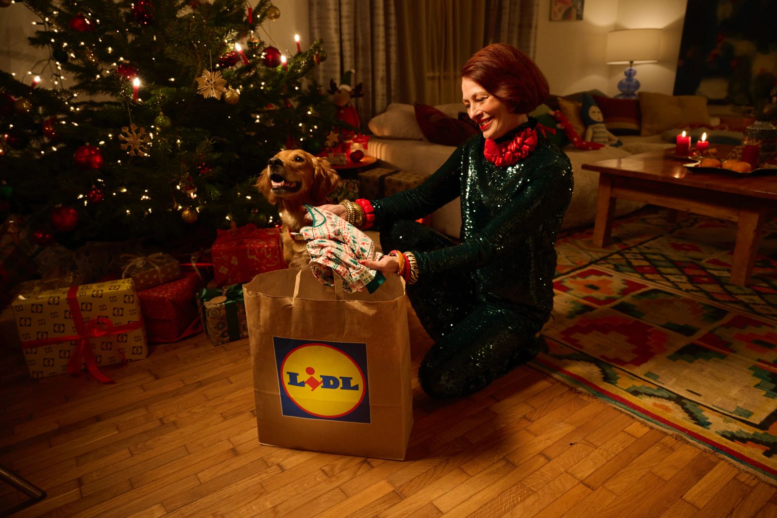 Eine Person in einem glitzernden grünen Outfit packt Geschenke aus einer Lidl-Tüte neben einem Hund und einem Weihnachtsbaum aus