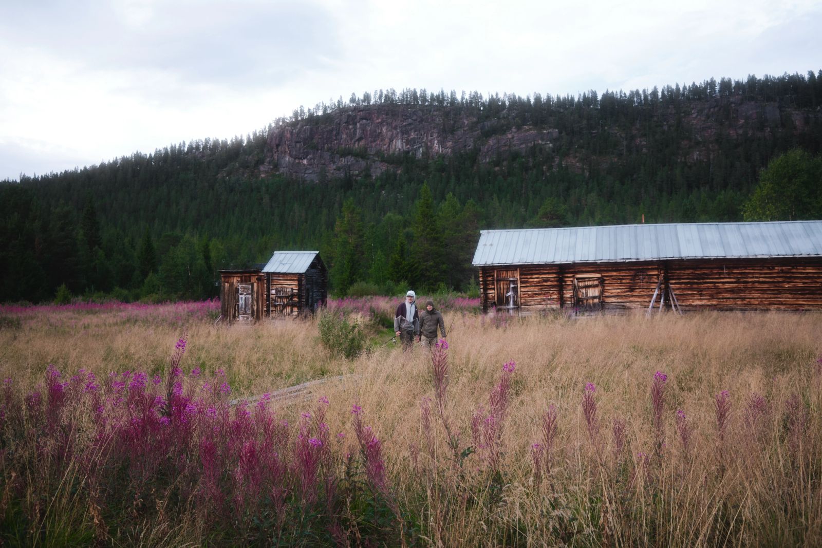 Zwei Wanderer vor traditionellen Holzhütten auf einer Blumenwiese in schwedischer Berglandschaft.