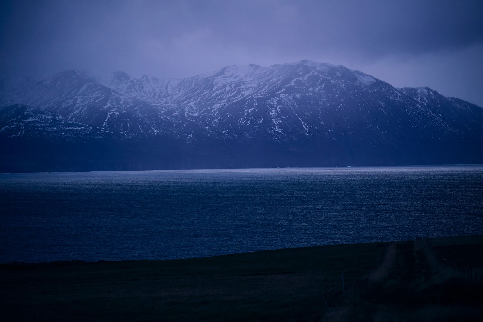 Eine weite isländische Küstenlandschaft in der Dämmerung mit tiefblauem Meer und schneebedeckten Bergen im Hintergrund, fotografiert von Karl Scholten.
