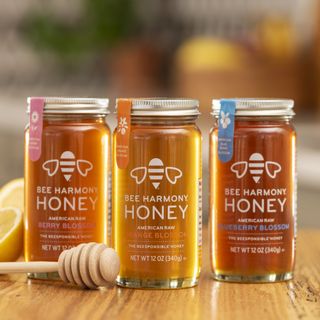 Three jars of Bee Harmony raw honey in Berry Blossom, Orange Blossom, and Blueberry Blossom flavors on a wooden table with a honey dipper.