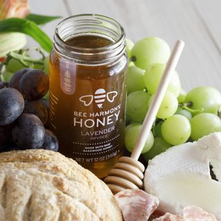Jar of Bee Harmony Lavender Infused Honey with grapes, bread, cheese, and a honey dipper on a table.