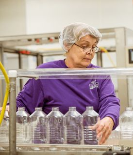 A worker wearing a hairnet and glasses inspects empty plastic bottles on a production line in a factory setting.