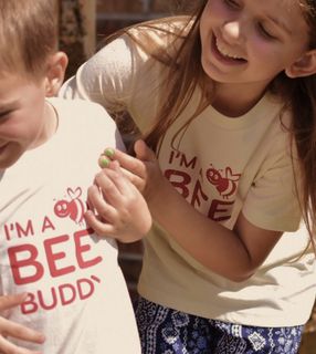 Two children in "I'm a bee budy" themed shirts, a boy and a girl, are smiling and playing.
