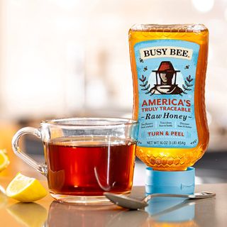 Clear glass mug with tea next to bottle of Busy Bee Raw Honey, lemon slice and spoon