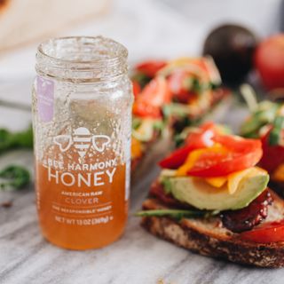 A half-empty jar of Bee Harmony American Raw Clover honey sits next to a toast topped with avocado, tomato, and peppers.
