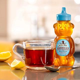 12 OZ Busy Bee Raw USA Honey bottle next to cup of tea and a spoon and a couple of lemon slices