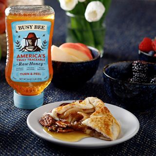 Busy bee honey squeezable bottle next to plate with a delicious pecans pastry cover of honey next to a couple of fruit plates with berries and apples