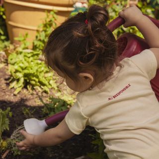 Toddler in a Beesponsible onesie watering a flower garden