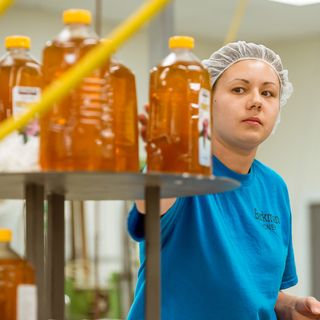 A worker wearing a hairnet and blue 'Barkman Honey' shirt inspects large bottles of honey on a production line in a factory setting.