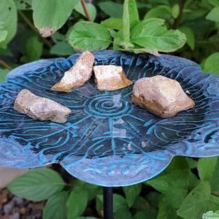A close-up of a blue glass birdbath filled with water and several rocks, surrounded by green leaves.