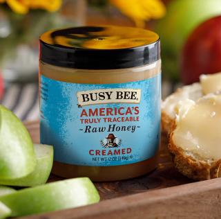 A jar of "Busy Bee Creamed Raw Honey" is on a wooden tray with green apple slices and honey-spread bread.