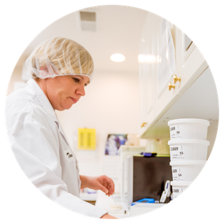 A lab technician wearing a hairnet and white coat works in a laboratory with labeled sample containers stacked on the counter.
