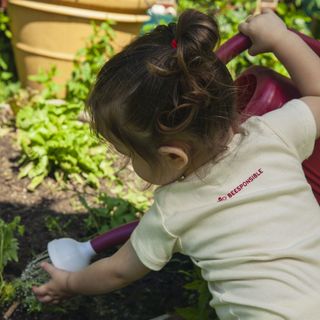 Toddler in a Beesponsible onesie watering a flower garden