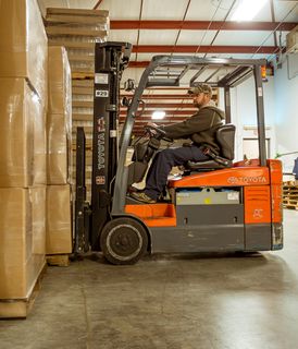 A worker operating a forklift to move stacks of large cardboard boxes inside a warehouse