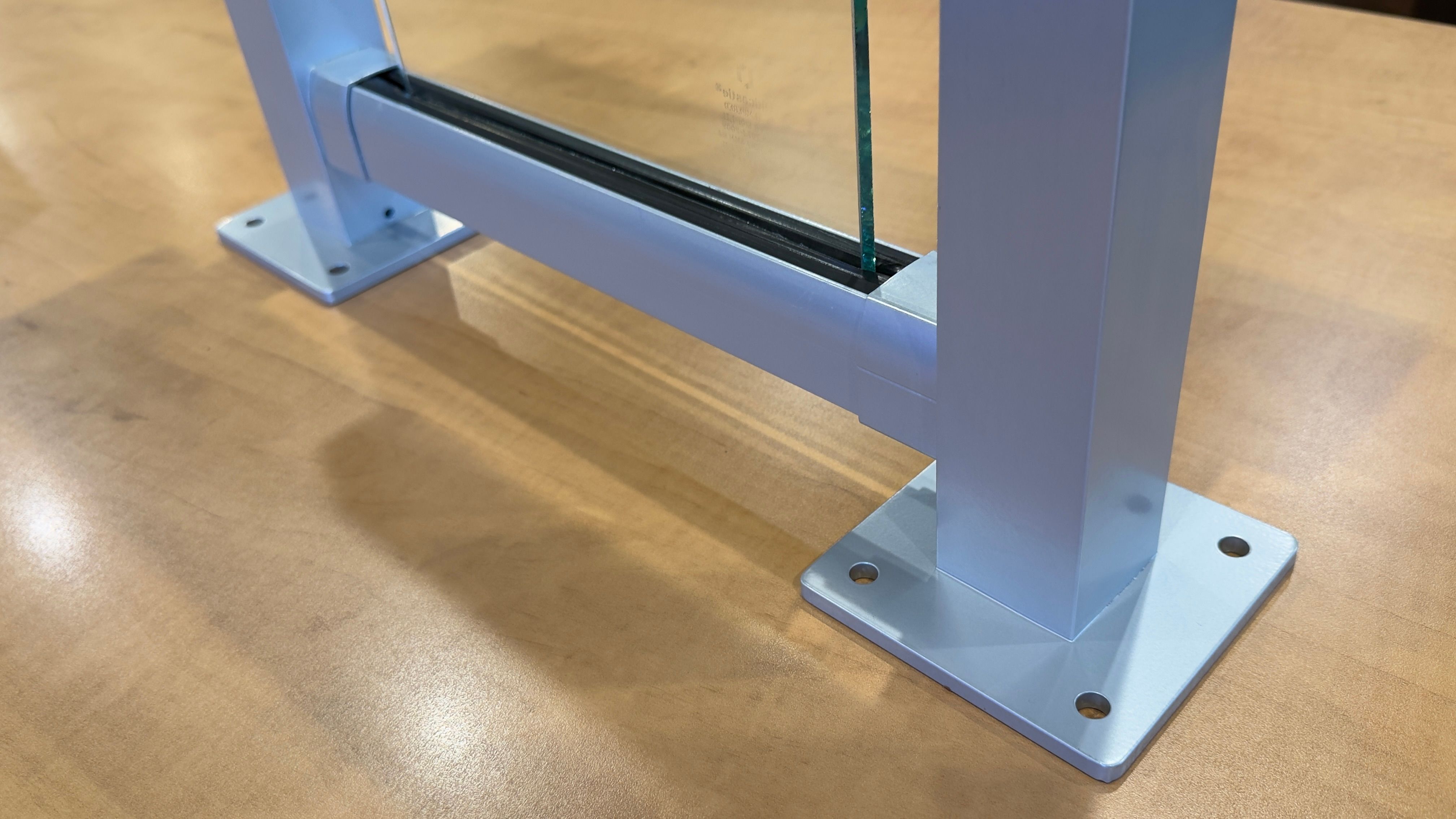 Close-up of glass railing mock-up showing surface mount base plates, bottom rail with rubber setting block, and tempered glass panel seated between two aluminum posts