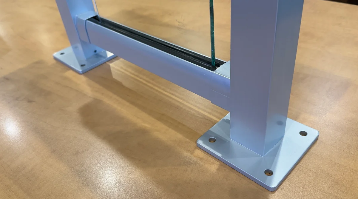 Close-up of glass railing mock-up showing surface mount base plates, bottom rail with rubber setting block, and tempered glass panel seated between two aluminum posts