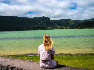 Vrouw zit met de rug naar de camera en kijkt uit over een groen meer met beboste heuvels.