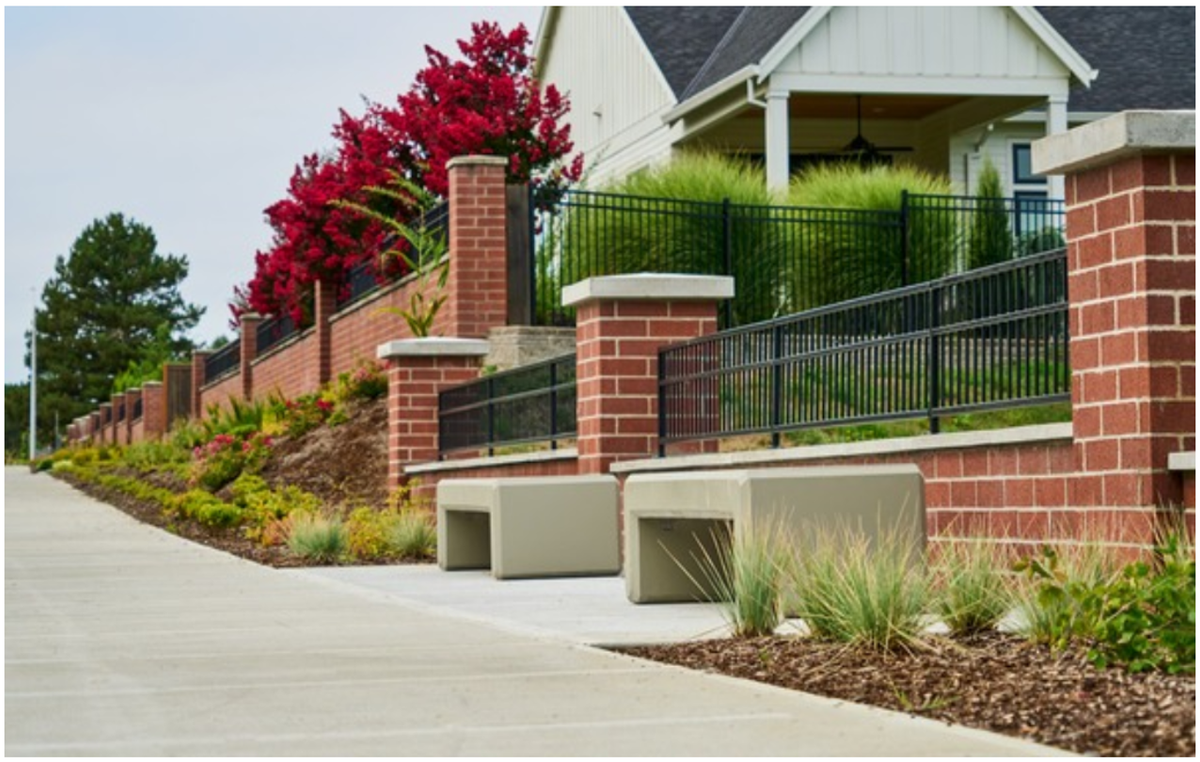 A concrete sidewalk runs along a brick retaining wall with black railings, two built-in concrete benches, and landscaping featuring red flowering bushes.