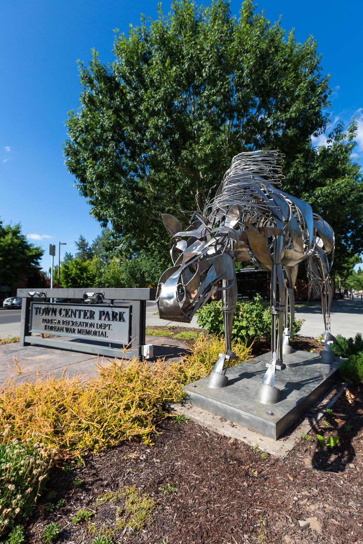 Large, segmented metal horse sculpture with a wire mane next to a sign for Town Center Park and Korean War Memorial.