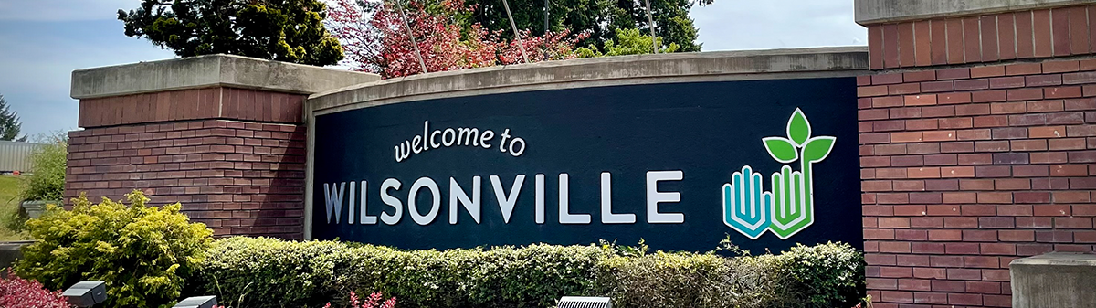 A "Welcome to Wilsonville" sign on a dark blue panel with white text and a green-blue logo, set in a brick structure with green bushes.