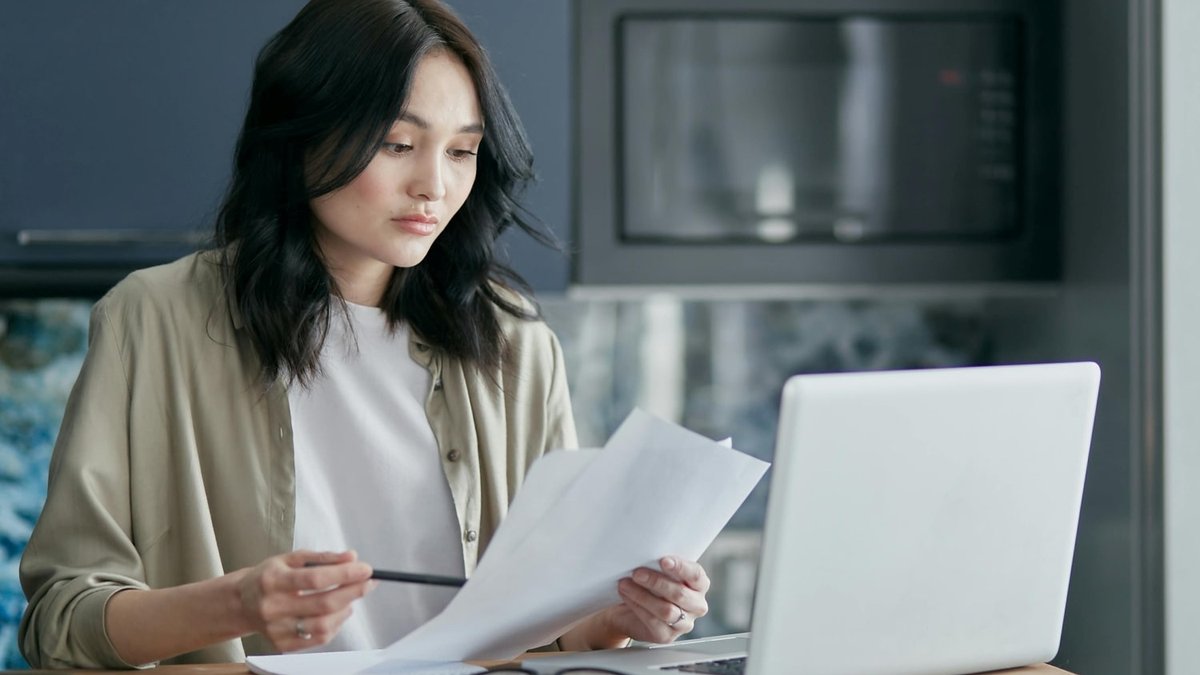 Woman looks at paperwork in front of a laptop.