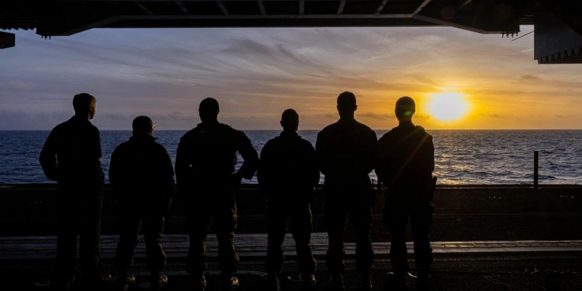 Silhouetted Navy sailors on aircraft carrier deck at sunset.