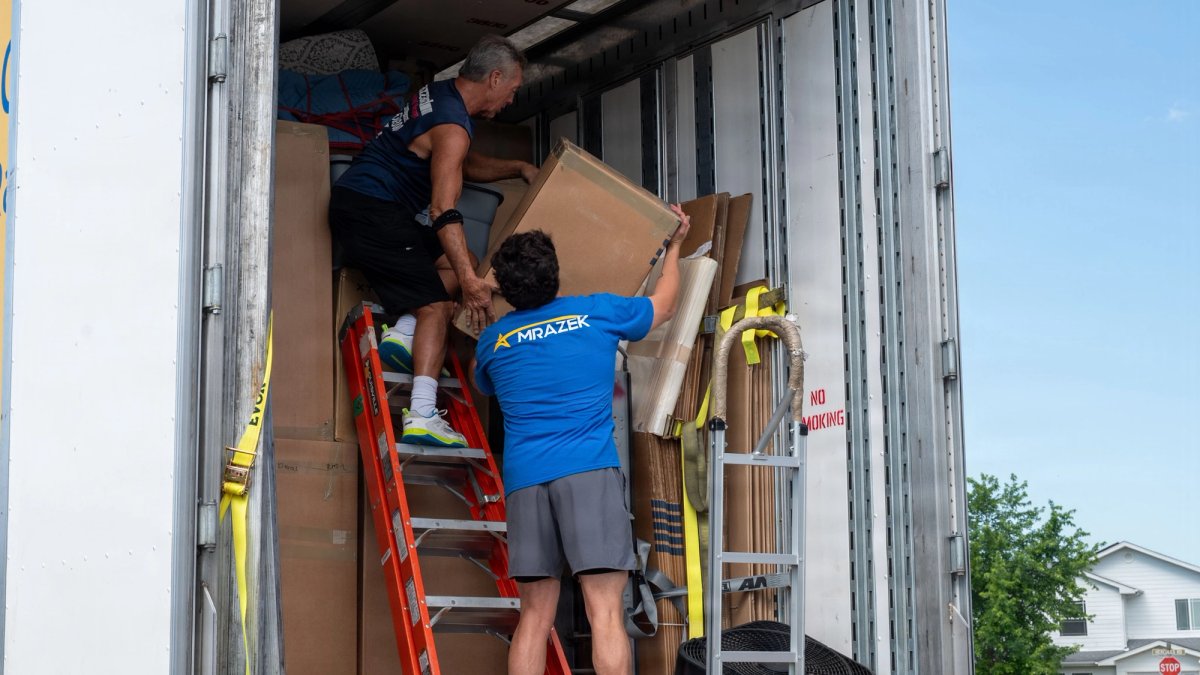 A mover hands a box to another standing on a ladder in a truck.