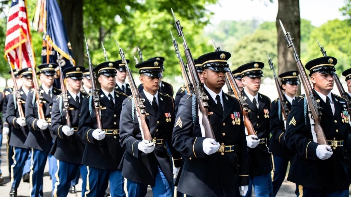 Army honor guard soldiers marching in formation with rifles and American flag during ceremony.