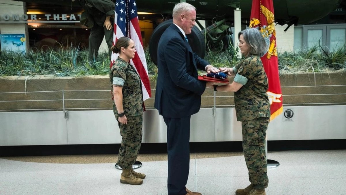 Civilian presenting folded flag to Marine Corps officer at military ceremony indoors
